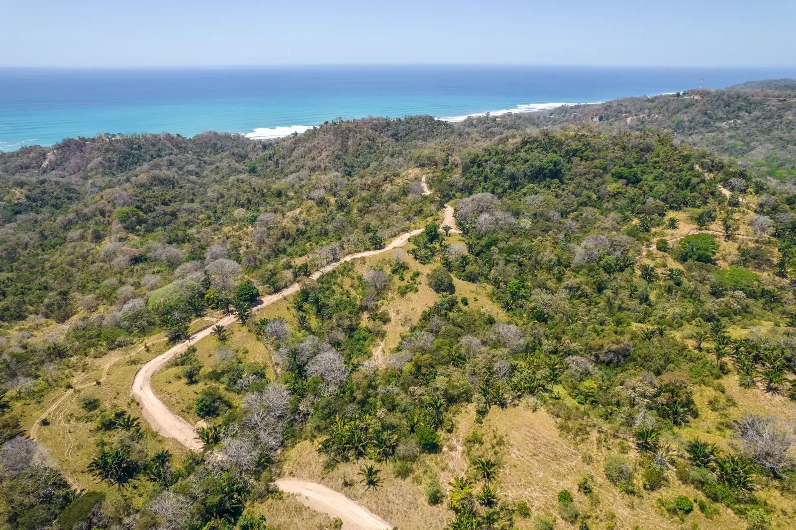 Aerial view of a dirt road winding across dry hills with the ocean beyond